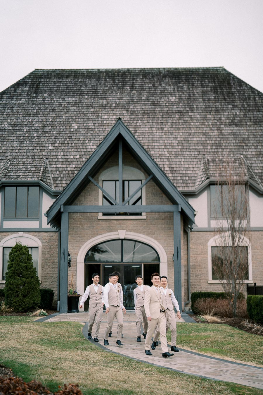 Groom and groomsmen in beige suits walking along a path in front of a large stone building with a steep roof, preparing for a wedding ceremony.
