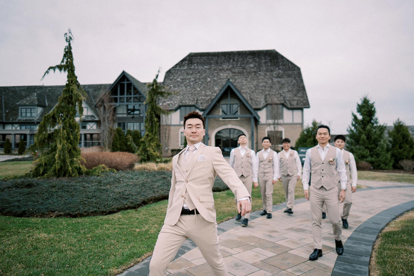 Groom and groomsmen in beige suits walking on a path in front of a picturesque estate.