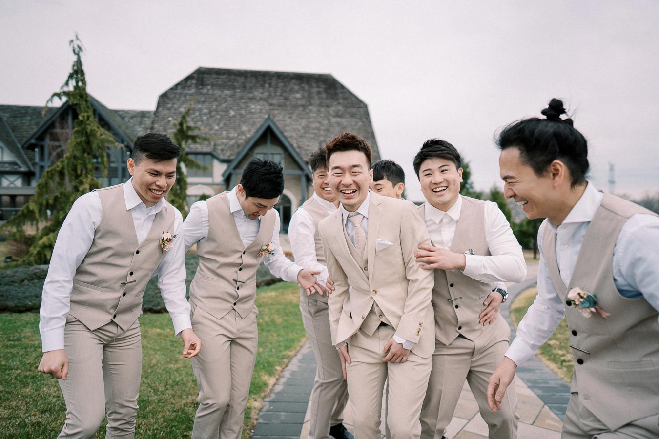 Groom and groomsmen laughing and walking together on a garden path, wearing beige suits and white shirts, in front of a rustic building.