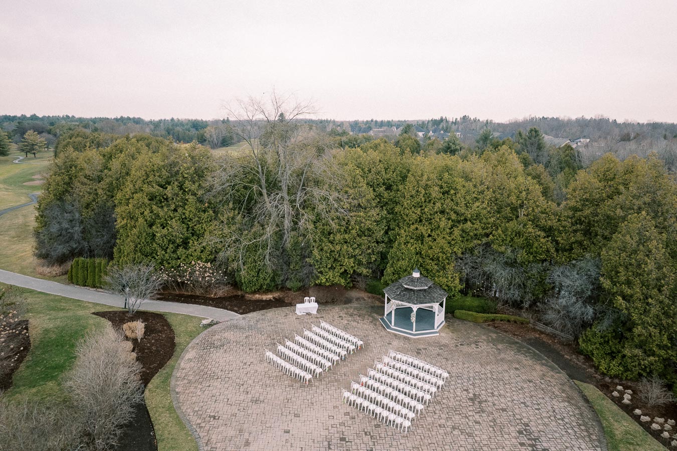 Aerial view of an outdoor wedding venue with rows of white chairs arranged on a paved area facing a charming gazebo, set amidst lush greenery and trees.