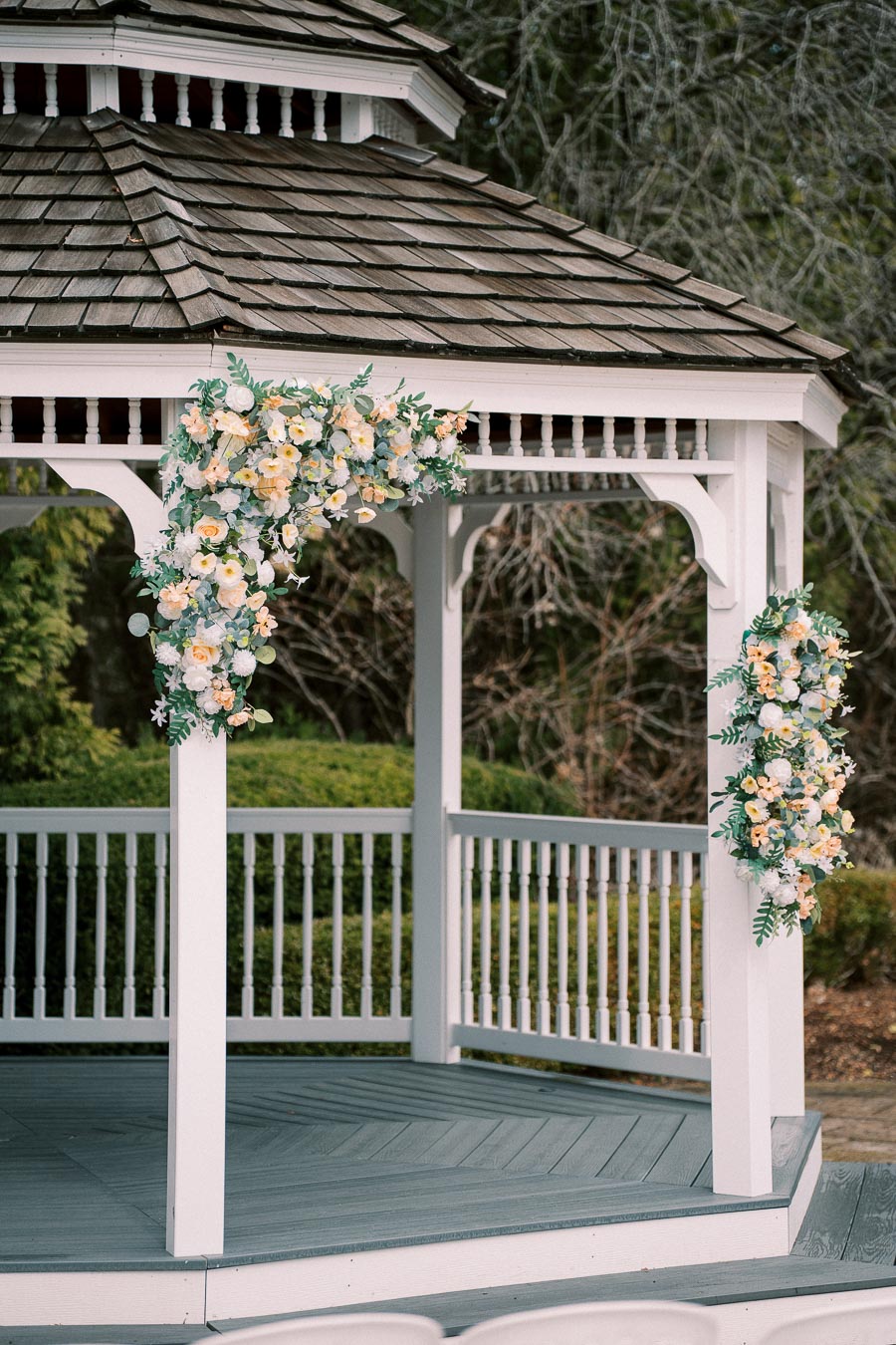 White wooden gazebo adorned with floral arrangements featuring white and yellow flowers, set against a backdrop of lush greenery. Ideal for outdoor weddings and garden events.