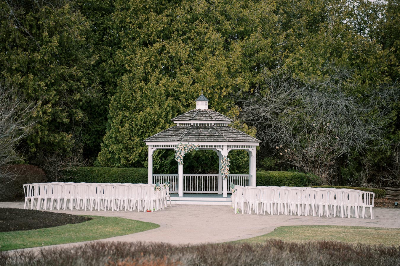 Outdoor wedding venue with a white gazebo and rows of chairs, surrounded by lush green trees.