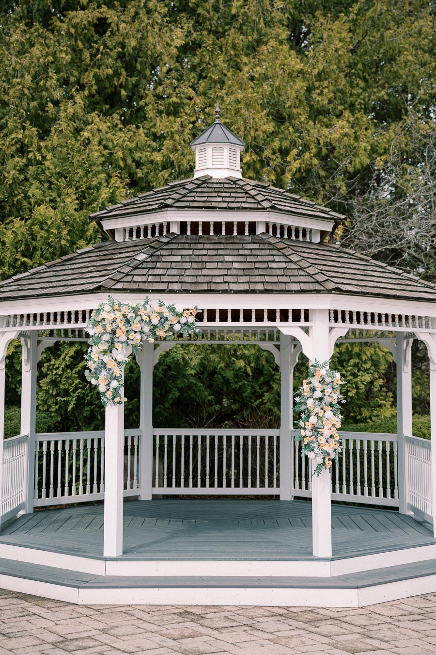 A charming white wooden gazebo adorned with floral arrangements stands on brick paving, set against lush green foliage, ideal for outdoor events or garden weddings.