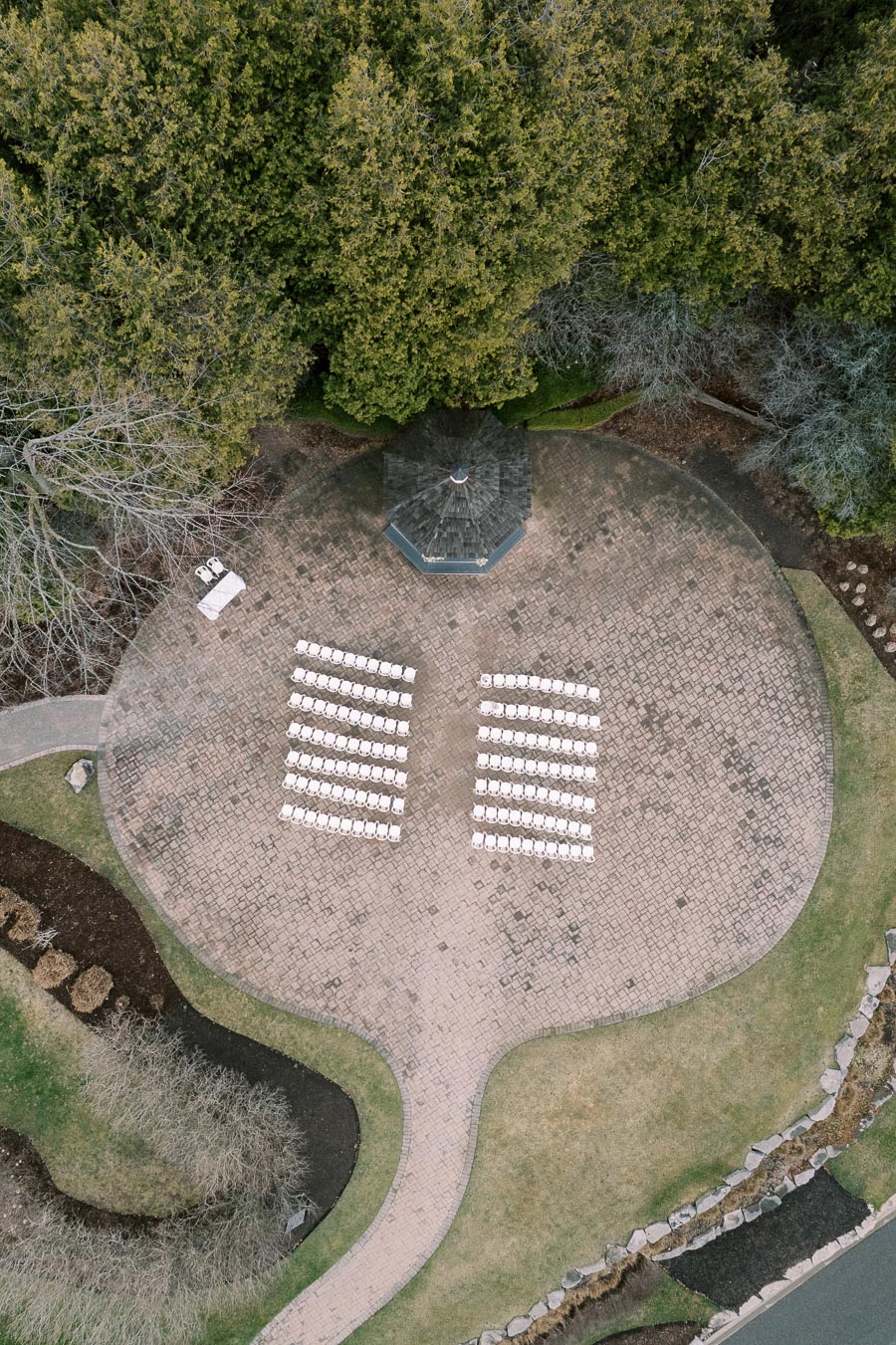 Aerial view of an outdoor event setup with rows of white chairs arranged on a paved area near a gazebo, surrounded by lush greenery and landscaping.