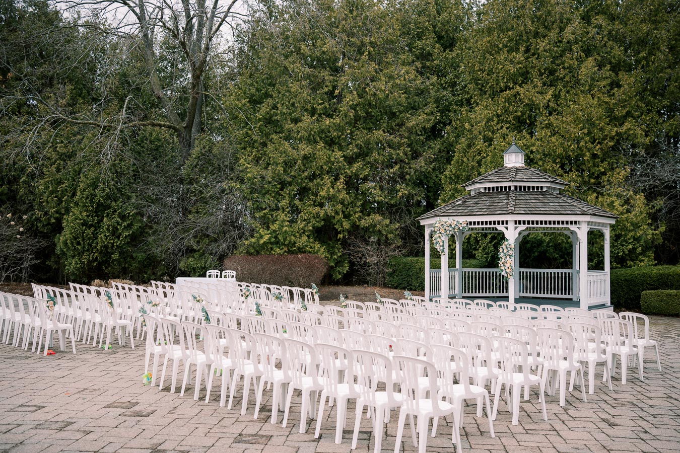Outdoor wedding venue with white chairs arranged in rows facing a decorated gazebo, surrounded by lush greenery.
