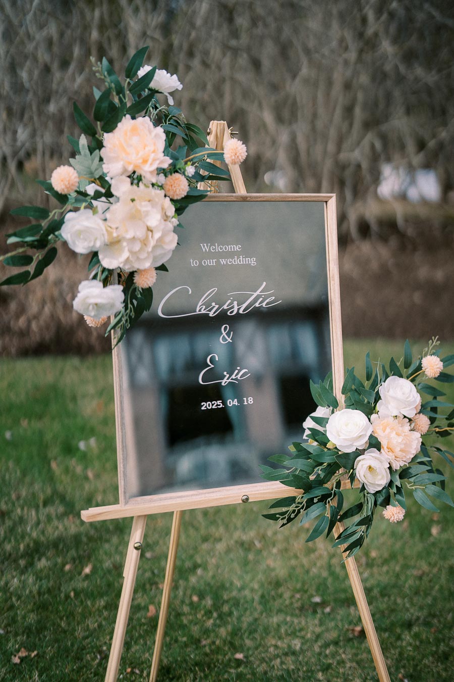 Elegant wedding welcome sign with white and peach flowers, displaying a couple's names and wedding date, set on a wooden easel in a garden setting.