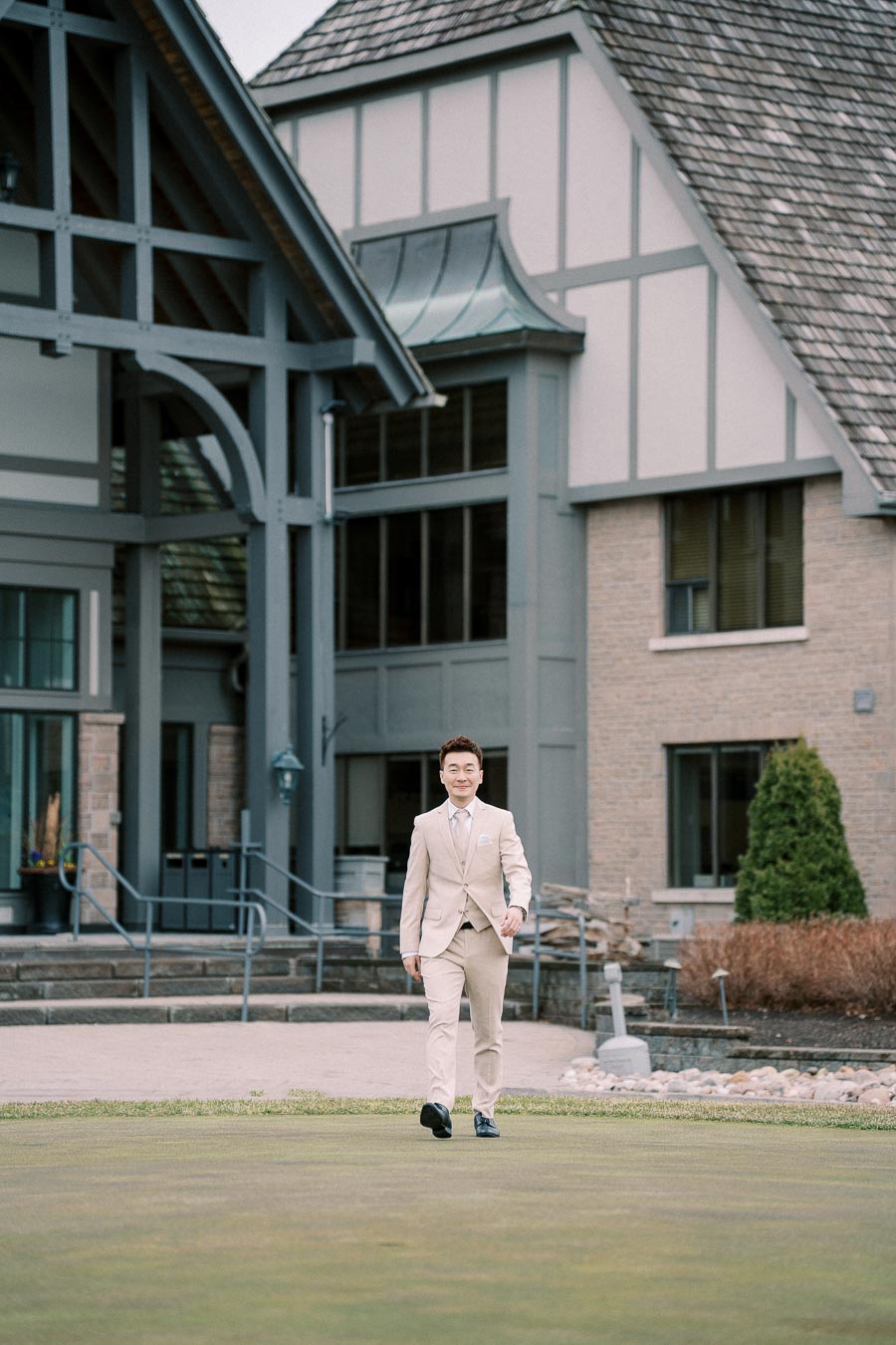 A man in a beige suit walking confidently in front of a traditional-style building with large windows and A-frame roof, surrounded by neatly kept landscaping.
