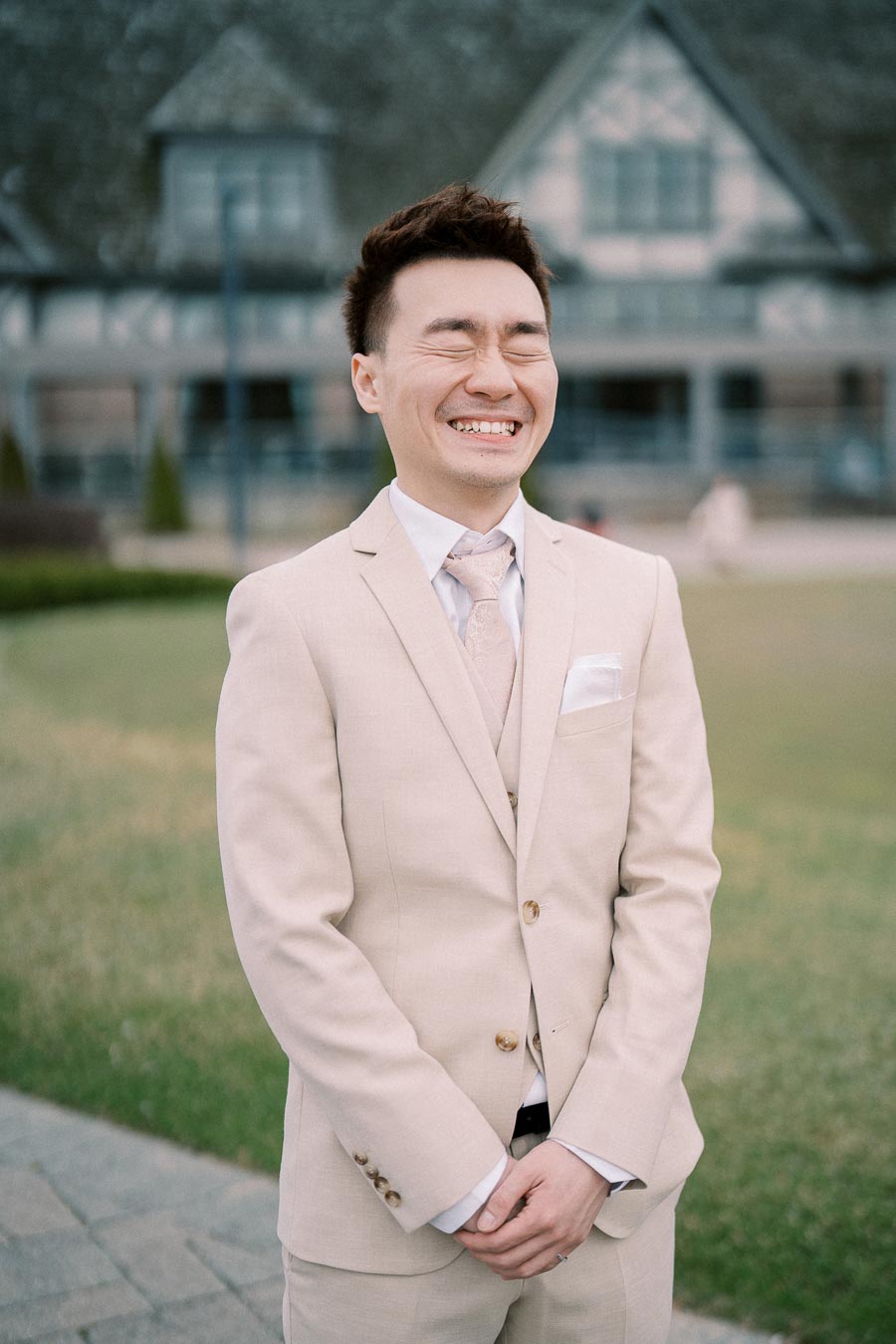 A person in a beige suit with a light-colored tie smiling broadly outside in front of a historic building with a lawn and pathway.