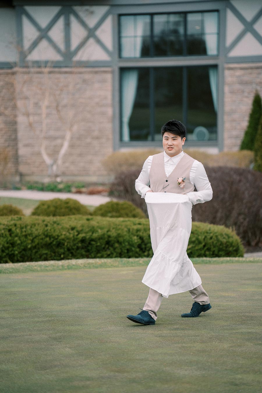 A person in a beige vest and white shirt gracefully walks on a well-manicured lawn, holding the hem of a long white garment with a backdrop of a building featuring large windows and green shrubbery.