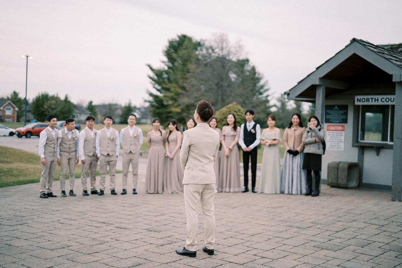 A group of people stand outdoors in formal attire near a small building labeled North Court, with a person in a beige suit facing them, likely during a wedding or formal event.