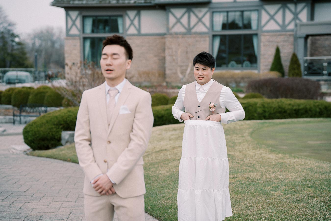 Two men in formal attire share a humorous moment outdoors; one in a beige suit with his eyes closed, and the other wearing a vest, holding up a white skirt with a playful expression, standing in front of a building amid green landscaping.