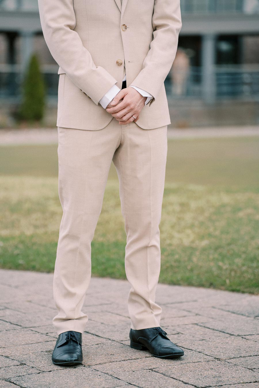Man in beige suit standing on a paved path outdoors, showcasing formal attire with black leather shoes.