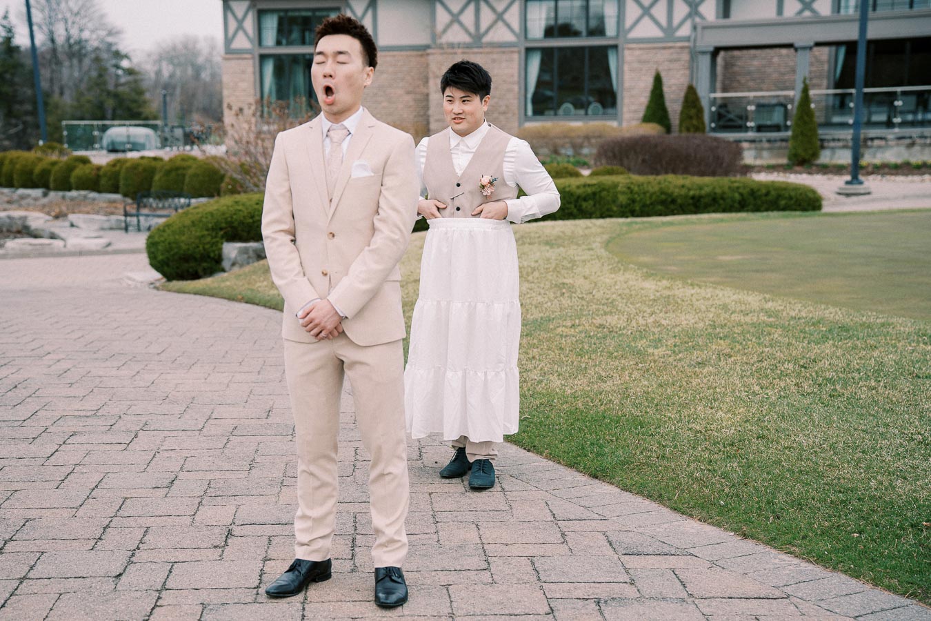 A person in a beige suit stands with eyes closed on a paved path, while another person playfully dresses in a white skirt and beige vest in the background, set in front of a building with landscaped greenery.