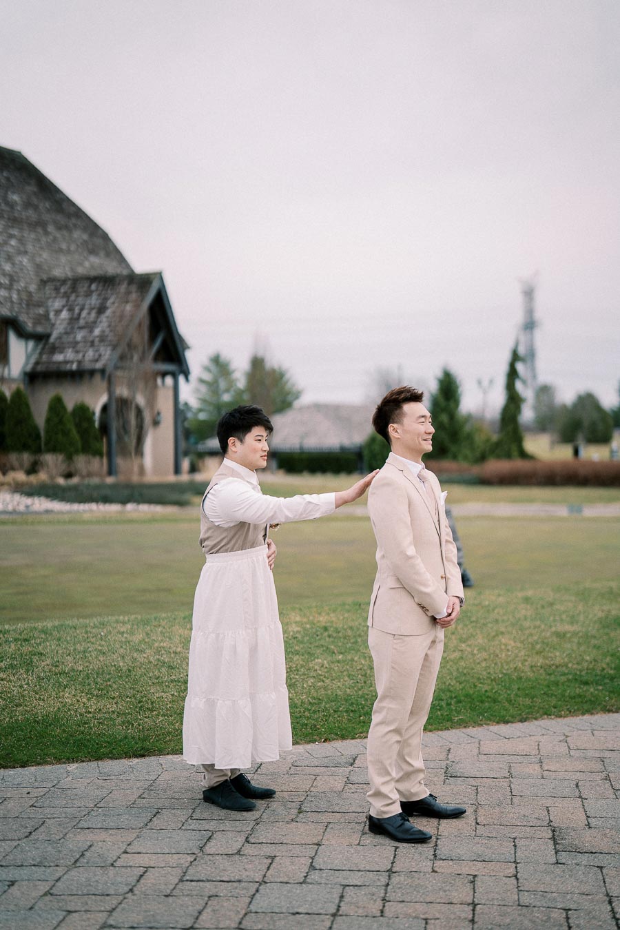 Two individuals sharing a joyful moment outdoors, with one wearing a suit and the other dressed in a long white dress. The scene is set against a backdrop of a rustic building and greenery, creating a serene and cheerful atmosphere.