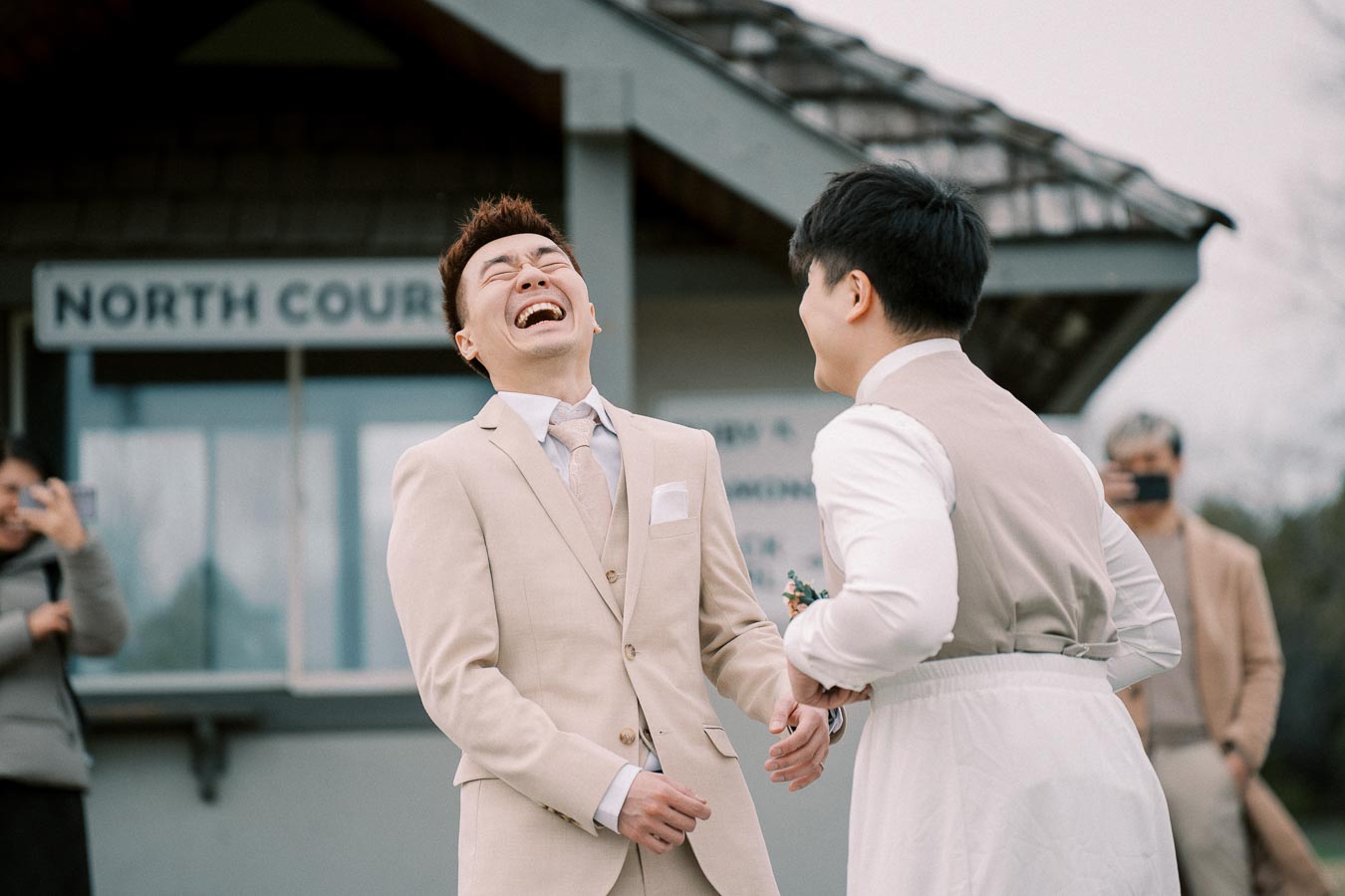 A joyful couple laughing and holding hands during an outdoor wedding ceremony, with guests capturing the moment in the background.
