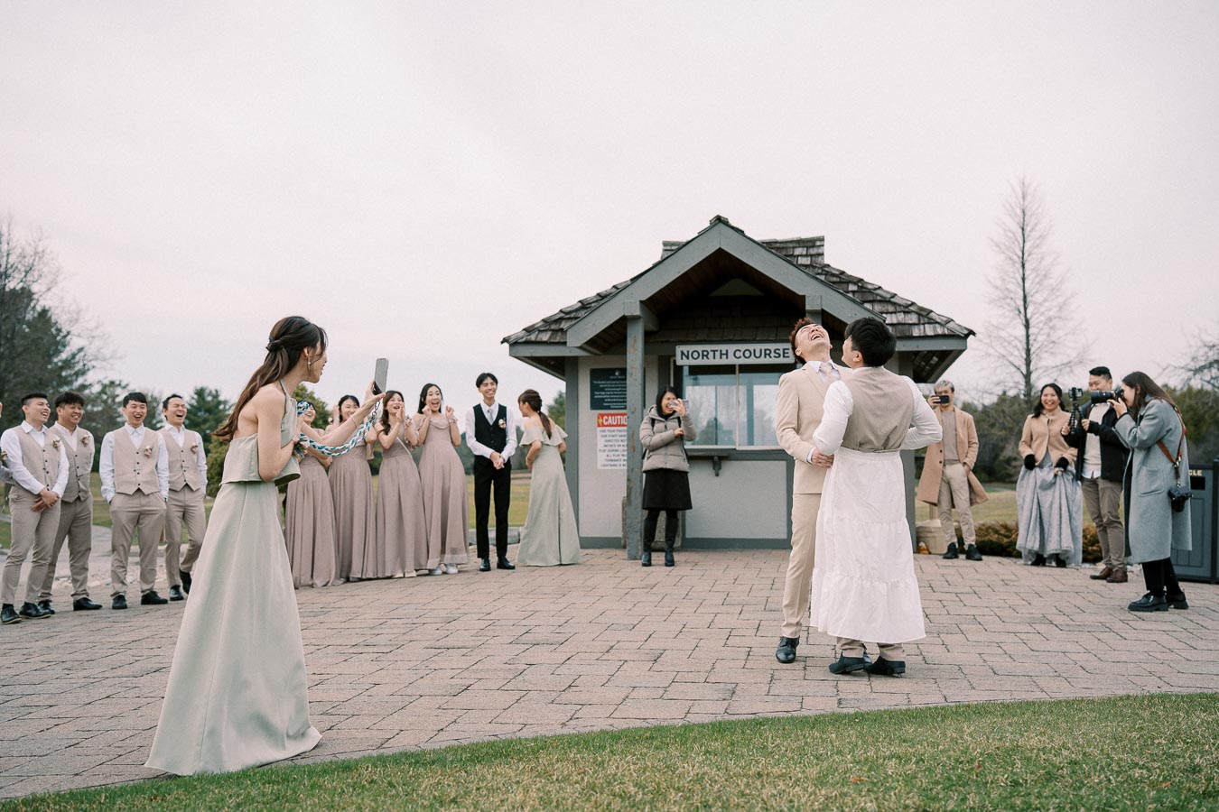 A joyful outdoor wedding ceremony with group posing, featuring grooms in traditional attire and bridesmaids in matching dresses, surrounded by guests and photographers capturing the moment.