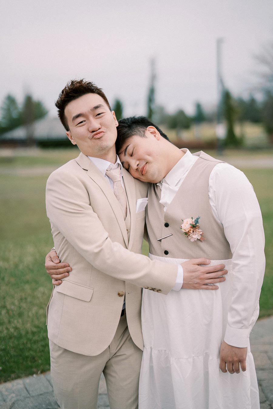 A joyful couple in elegant beige suits embrace outdoors, with one person playfully making a duck face while the other rests their head affectionately on their shoulder, surrounded by a serene, blurred green and gray landscape.