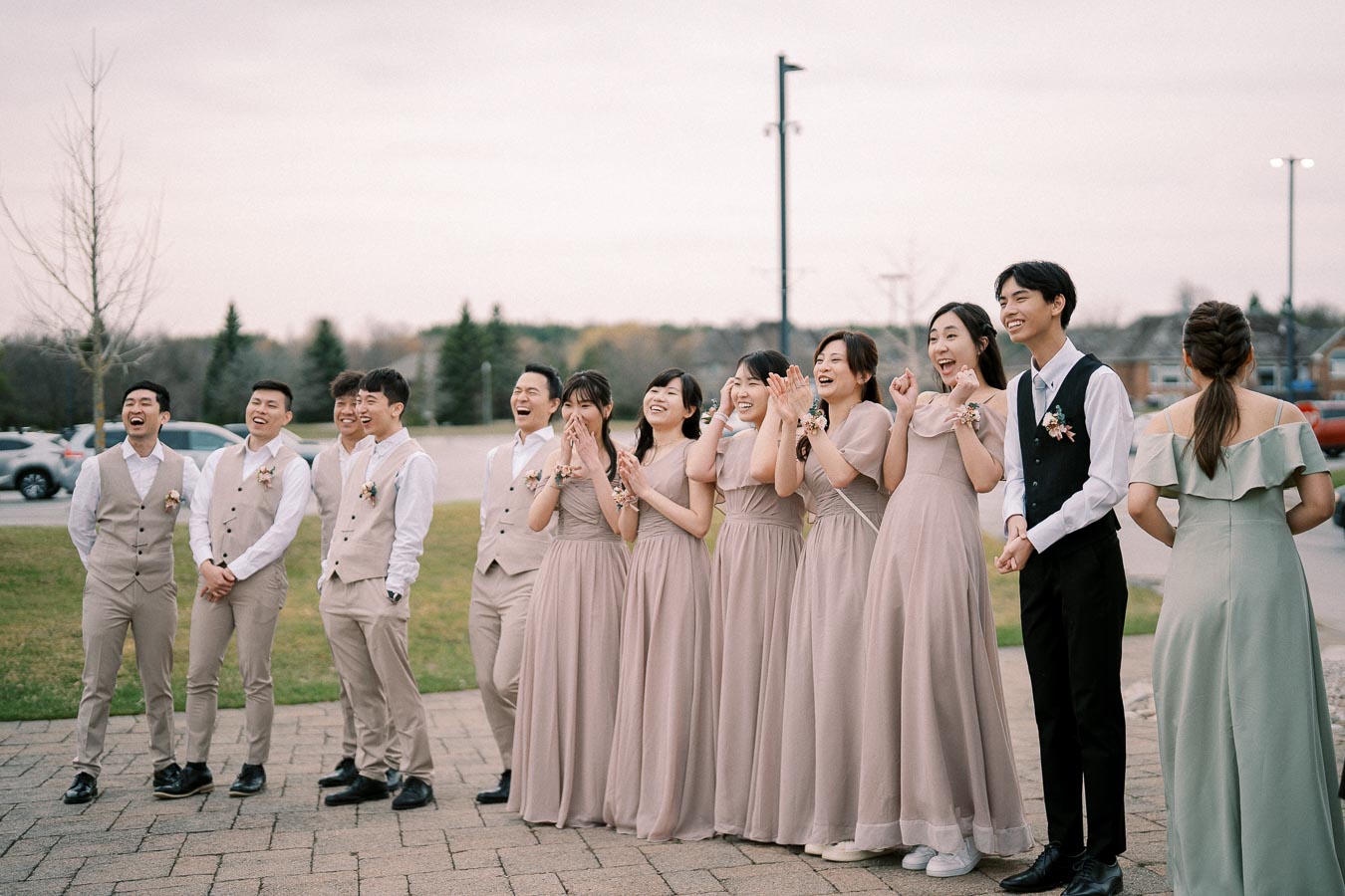 A group of bridesmaids and groomsmen laughing joyfully in a wedding celebration, wearing coordinated light beige and gray attire, standing outside on a paved area near greenery and trees, capturing a candid and cheerful moment.