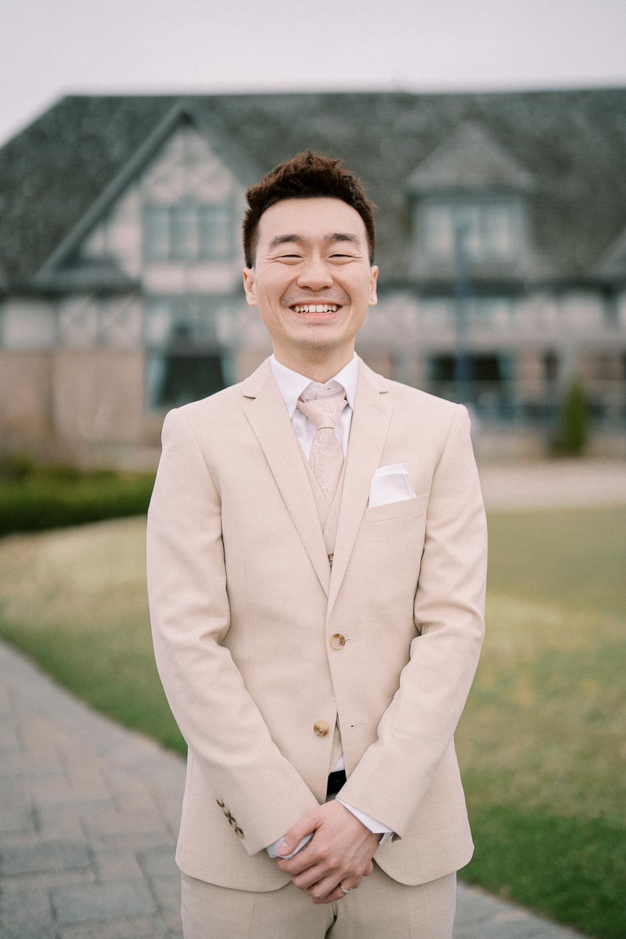 A man in a beige suit smiling warmly in front of a rustic building, standing on a stone pathway with greenery in the background.