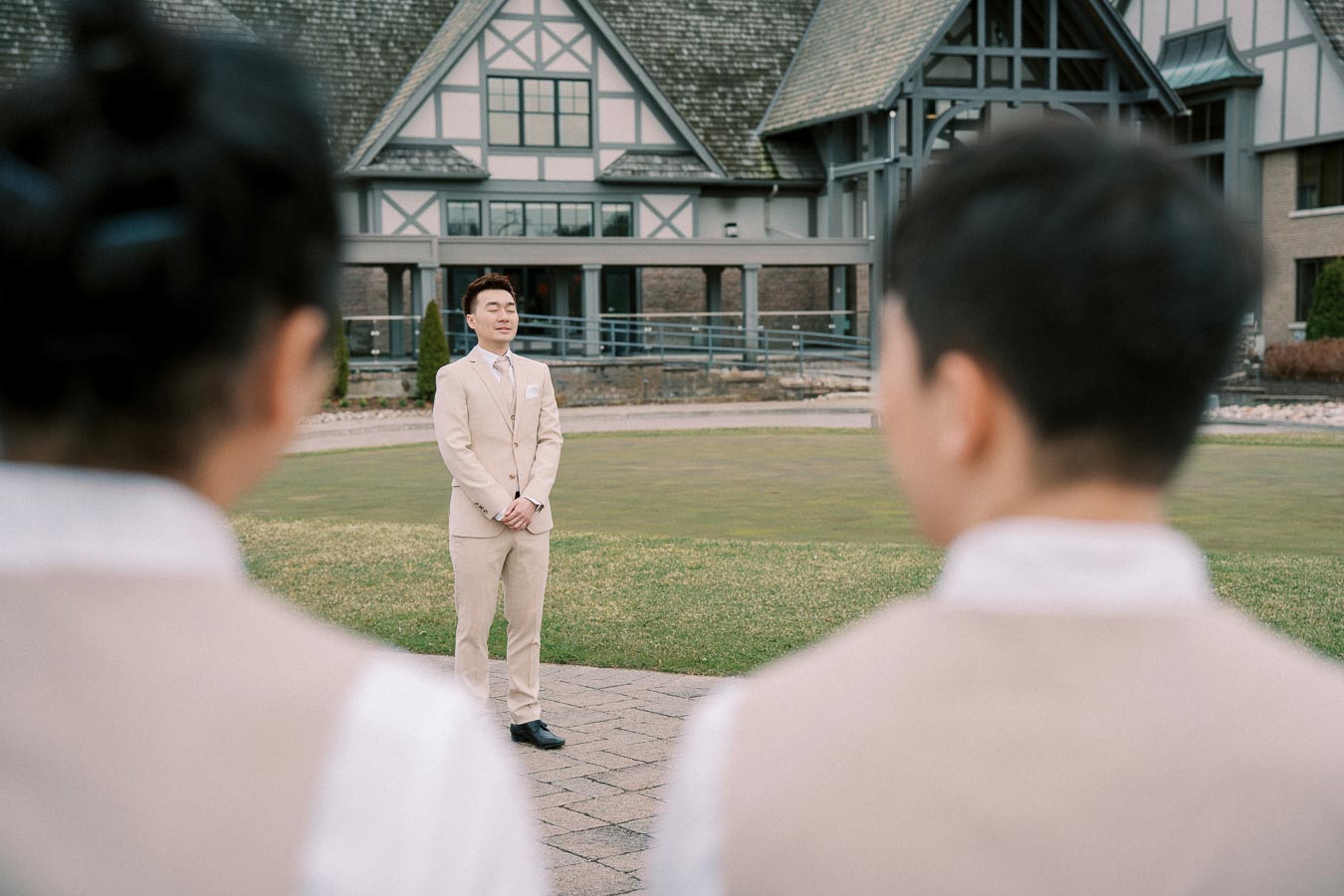 A groom in a beige suit stands smiling in front of a tudor-style building, with two blurred figures in the foreground during an outdoor wedding ceremony.