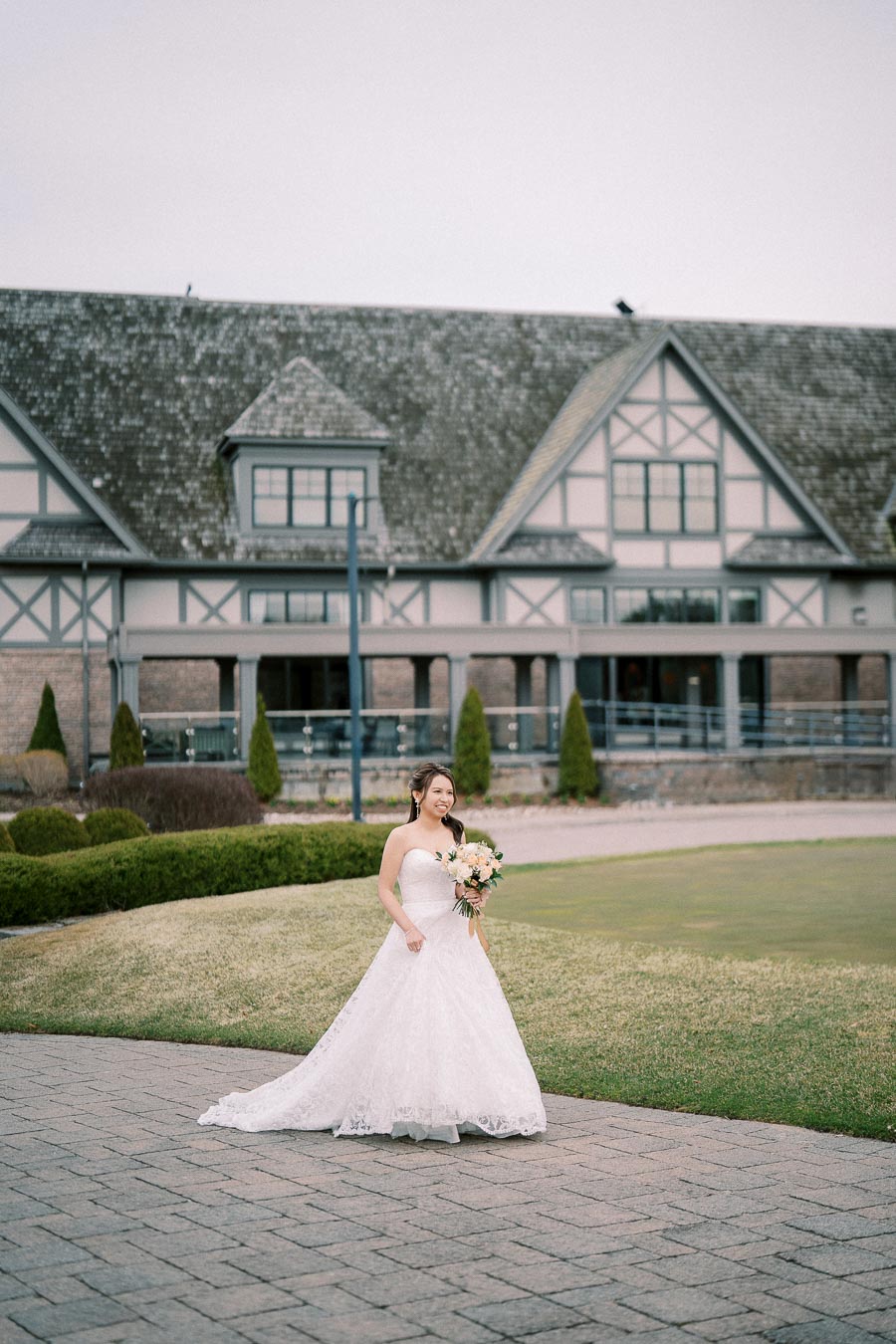 A bride in a white wedding gown holds a bouquet while standing on a paved walkway with a beautiful country house in the background, surrounded by greenery.