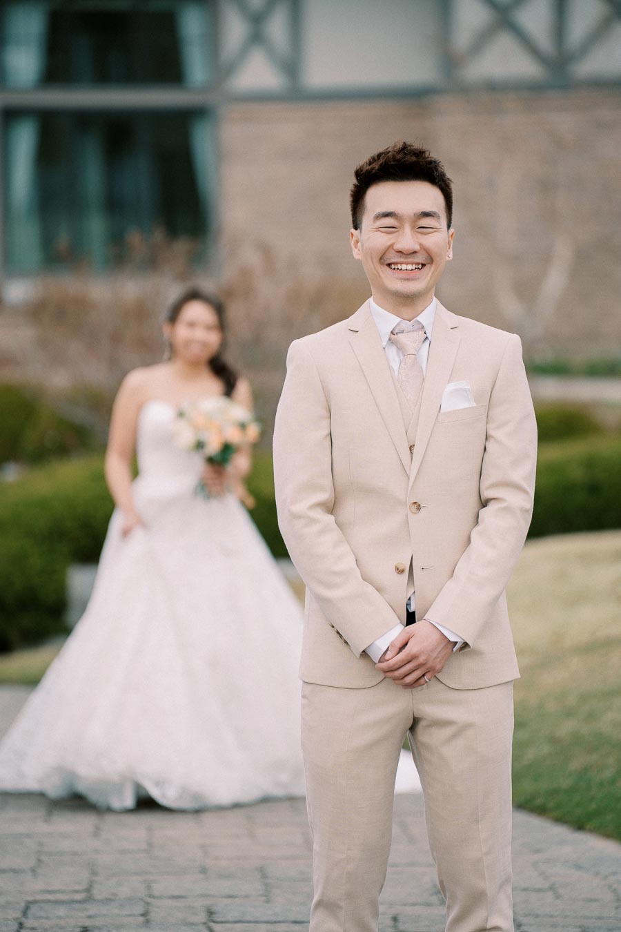 A groom in a light beige suit smiling joyfully with a bride in a white wedding dress holding a bouquet in the background, captured in an outdoor setting.
