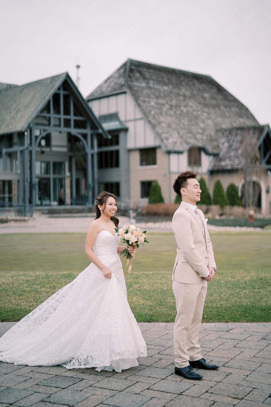 A bride in a white lace gown holding a bouquet walks towards a groom in a light beige suit, standing in front of a rustic stone building with a gabled roof, set on a green lawn.
