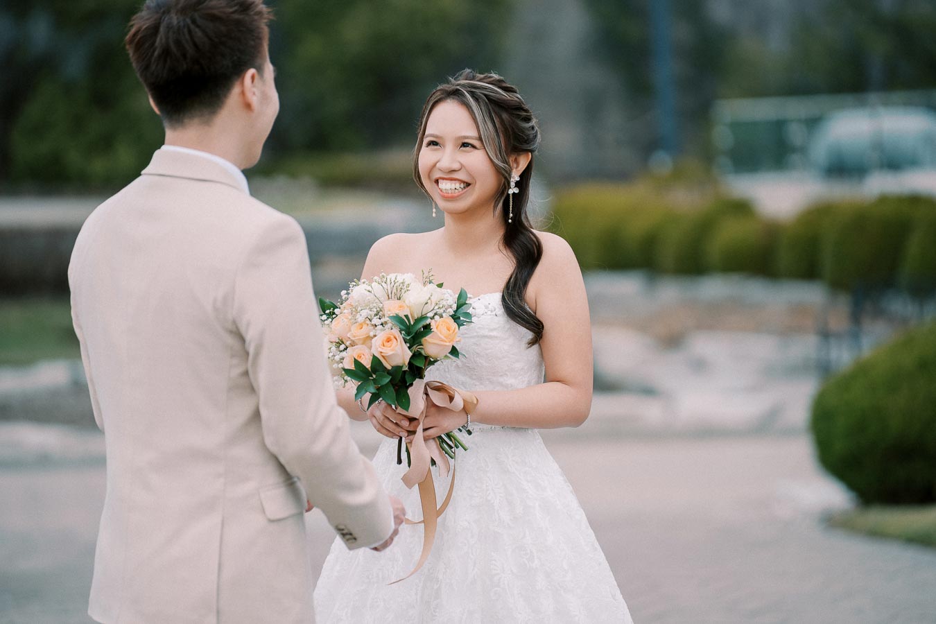 Smiling bride holding a bouquet of peach and white flowers faces a groom in a beige suit outdoors, showcasing a joyful wedding moment in a garden setting.