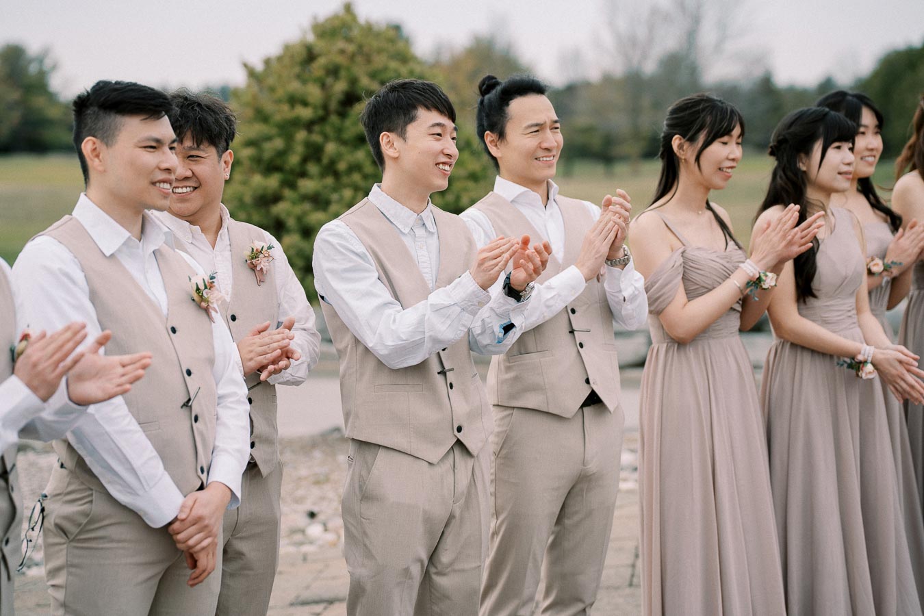 A group of bridesmaids and groomsmen in coordinated beige attire stand outdoors, clapping and smiling during a wedding ceremony.
