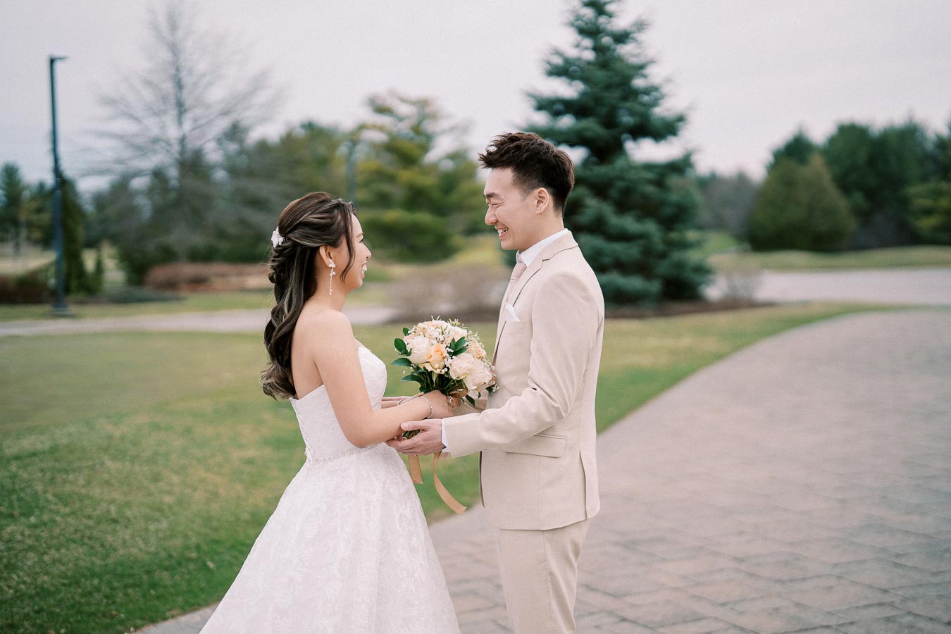 A bride and groom stand face-to-face outdoors, holding hands, with the bride wearing a white lace gown and the groom in a beige suit, surrounded by greenery and a paved path.