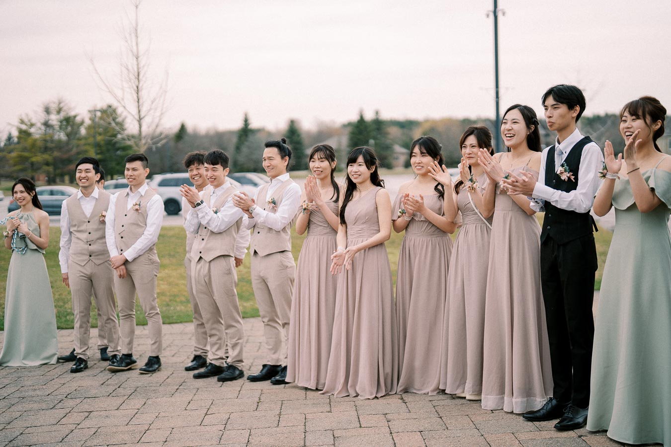 A joyful group of bridesmaids and groomsmen in matching beige outfits and elegant dresses, clapping and smiling at an outdoor wedding ceremony.