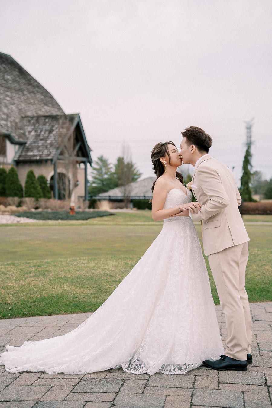 A bride and groom share a romantic kiss outside a rustic venue, with the bride in a lace wedding gown and the groom in a beige suit, capturing a beautiful wedding moment.