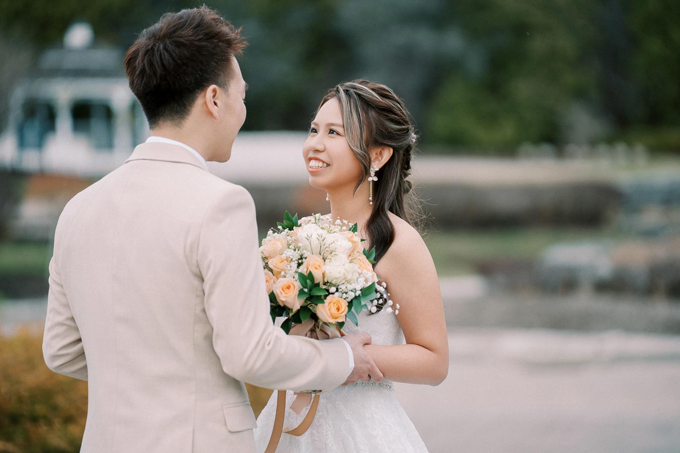 A bride and groom smiling at each other during an outdoor wedding ceremony, with the bride holding a bouquet of peach and white flowers, creating a romantic and happy atmosphere.