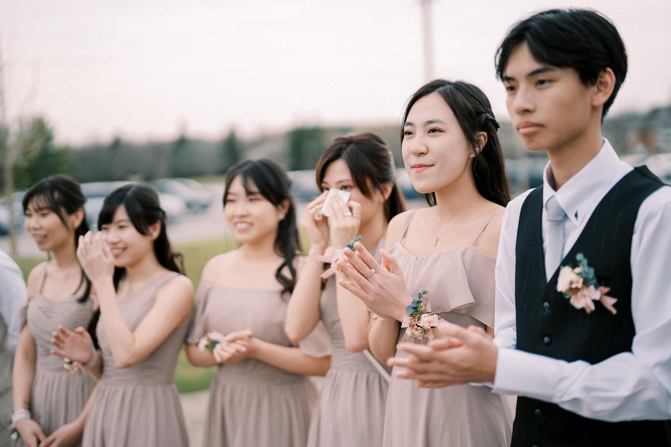 Young wedding guests in formal attire clap and smile outdoors, with bridesmaids in matching beige dresses and floral wristbands.