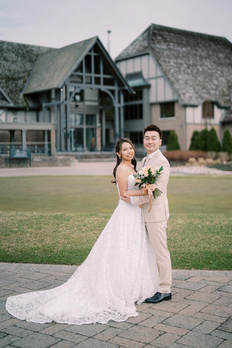 A couple in wedding attire poses happily in front of a rustic building, with the groom in a beige suit holding the bride, who is wearing a white lace gown, while standing on a stone pathway with greenery in the background.
