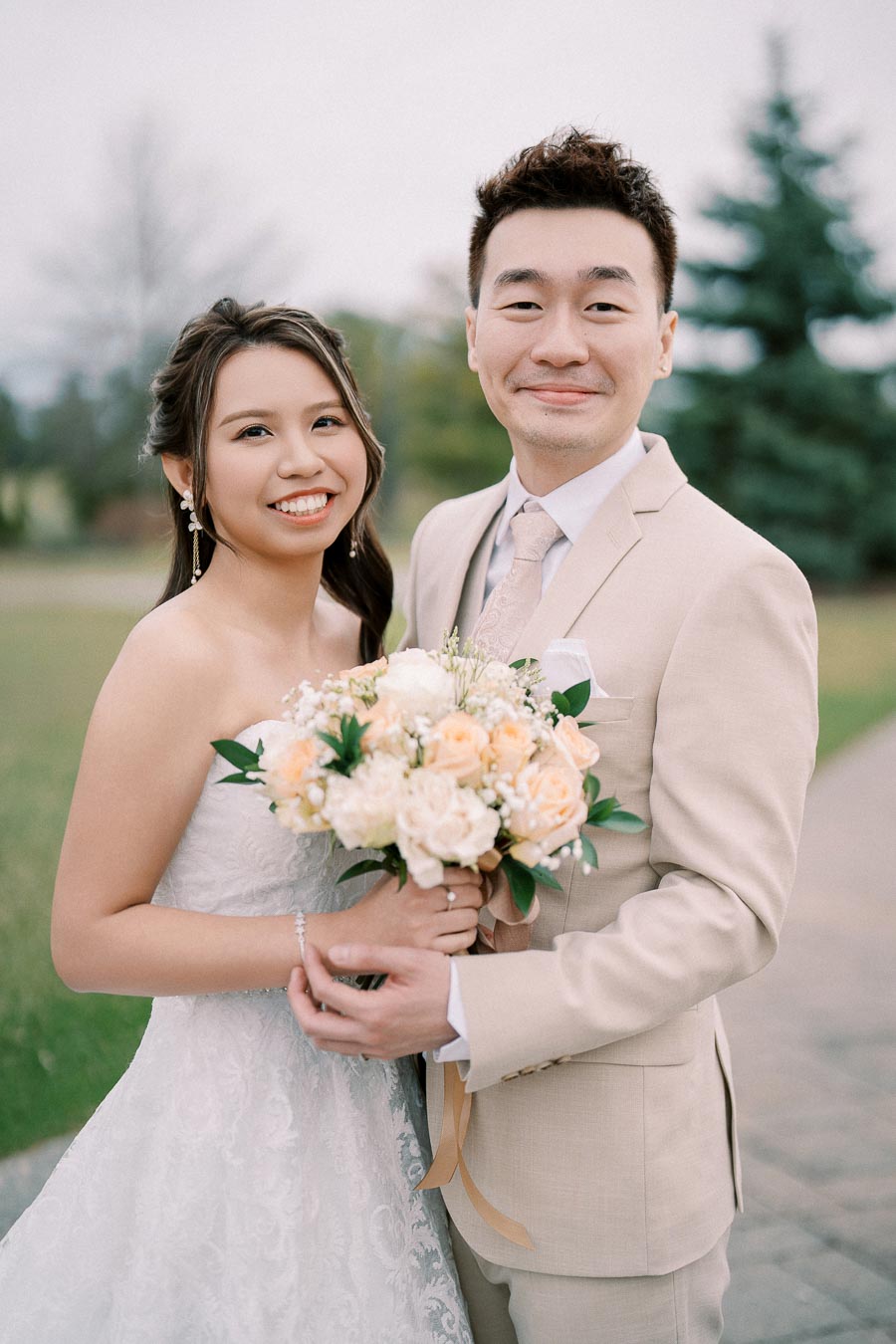 A happy bride and groom pose outdoors in a park setting, with the bride holding a bouquet of pastel roses and greenery. The groom is wearing a beige suit, and the bride is in a white lace wedding dress, both smiling warmly.