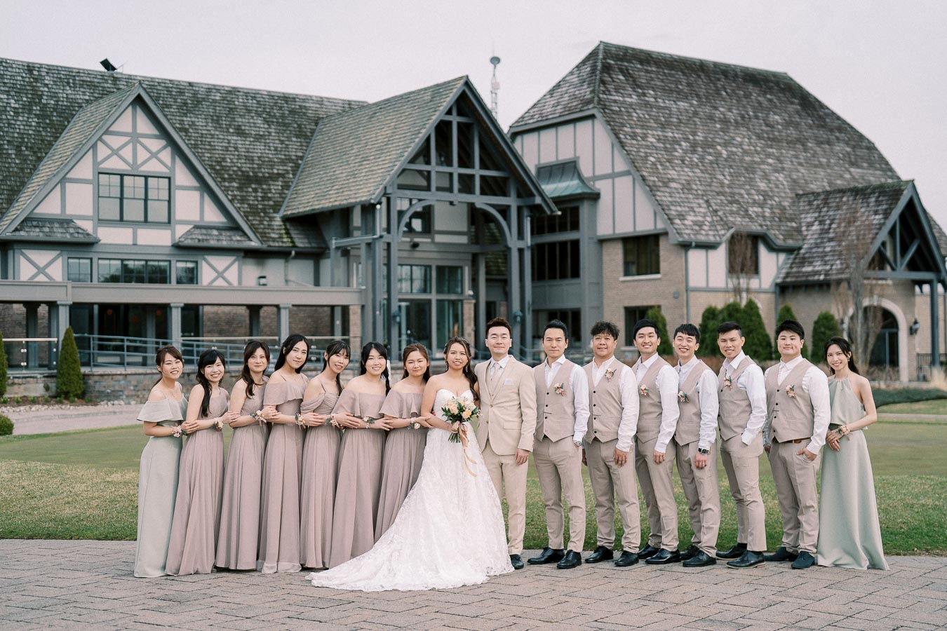 A wedding party poses in front of a rustic, Tudor-style mansion. The bride, in a white gown, and the groom, in a beige suit, stand at the center, surrounded by bridesmaids in matching taupe dresses and groomsmen in beige vests, all smiling warmly on a landscaped lawn.