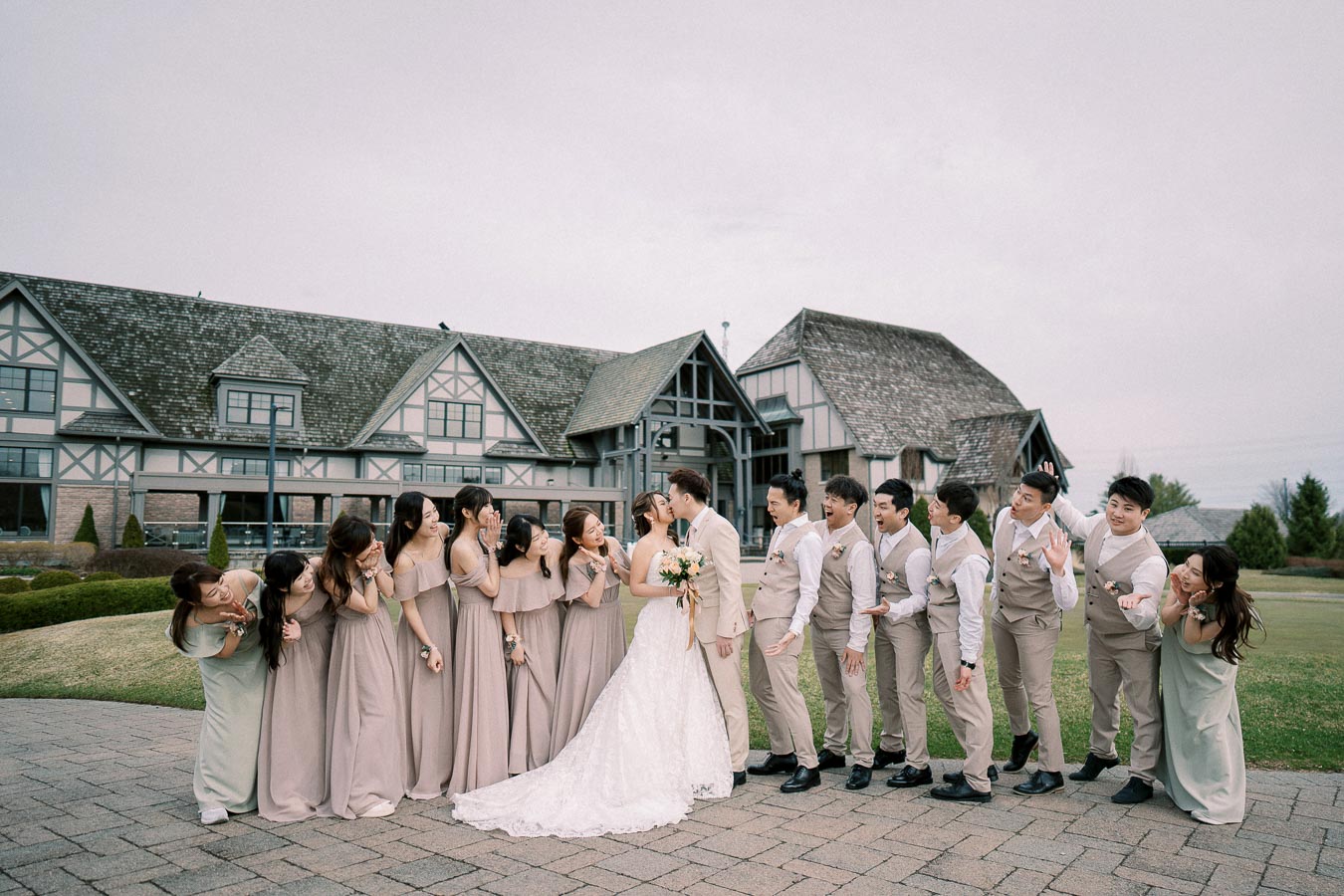 Wedding party celebrating at a charming venue, featuring a bride in a white gown and groom in a light suit kissing, surrounded by bridesmaids in elegant beige dresses and groomsmen in matching vests. Tudor-style building in the background.