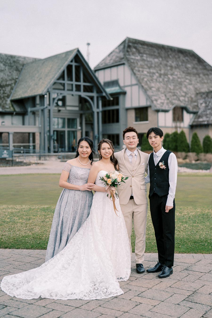 A wedding couple posing with two guests in elegant attire, standing on a paved path in front of a classic, rustic building with a lawn and green surroundings. The bride is in a white lace gown, holding a bouquet, and the groom is in a beige suit.