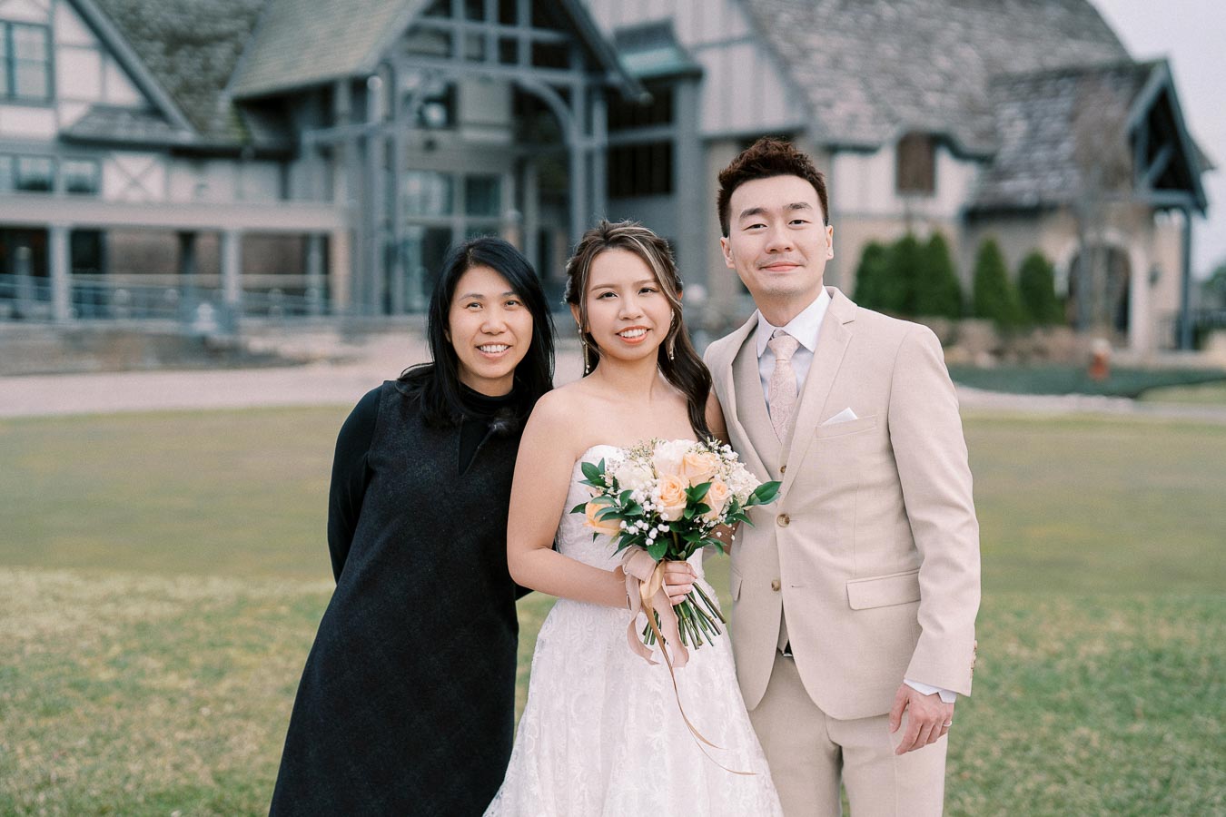 A wedding couple stands smiling with a friend in front of a rustic building, the bride holding a bouquet of white and peach flowers, in an outdoor setting.