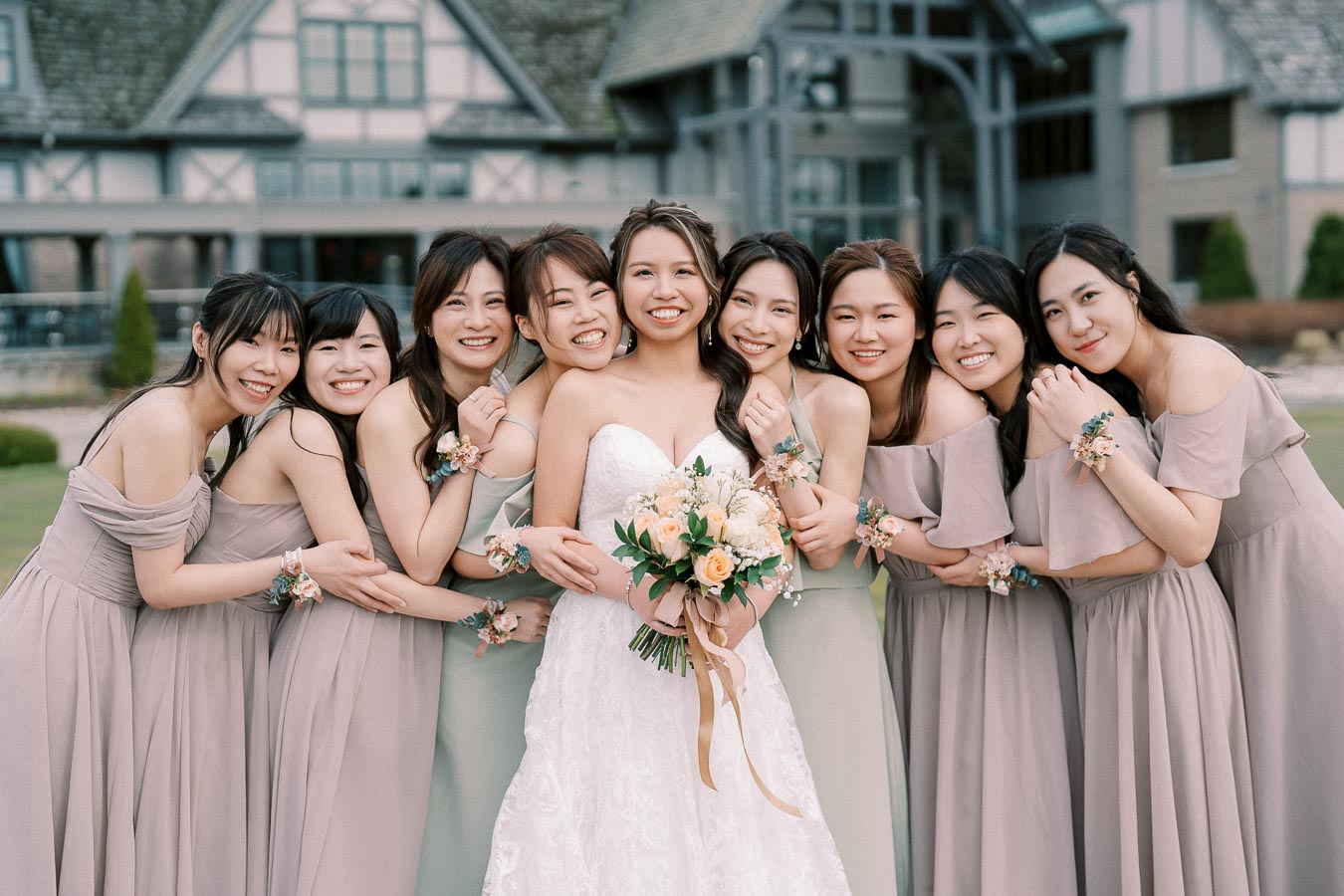 A joyful bride in a white lace wedding dress holding a bouquet of peach flowers, surrounded by her eight bridesmaids in matching gray dresses, all smiling and embracing outdoors in front of a rustic building.