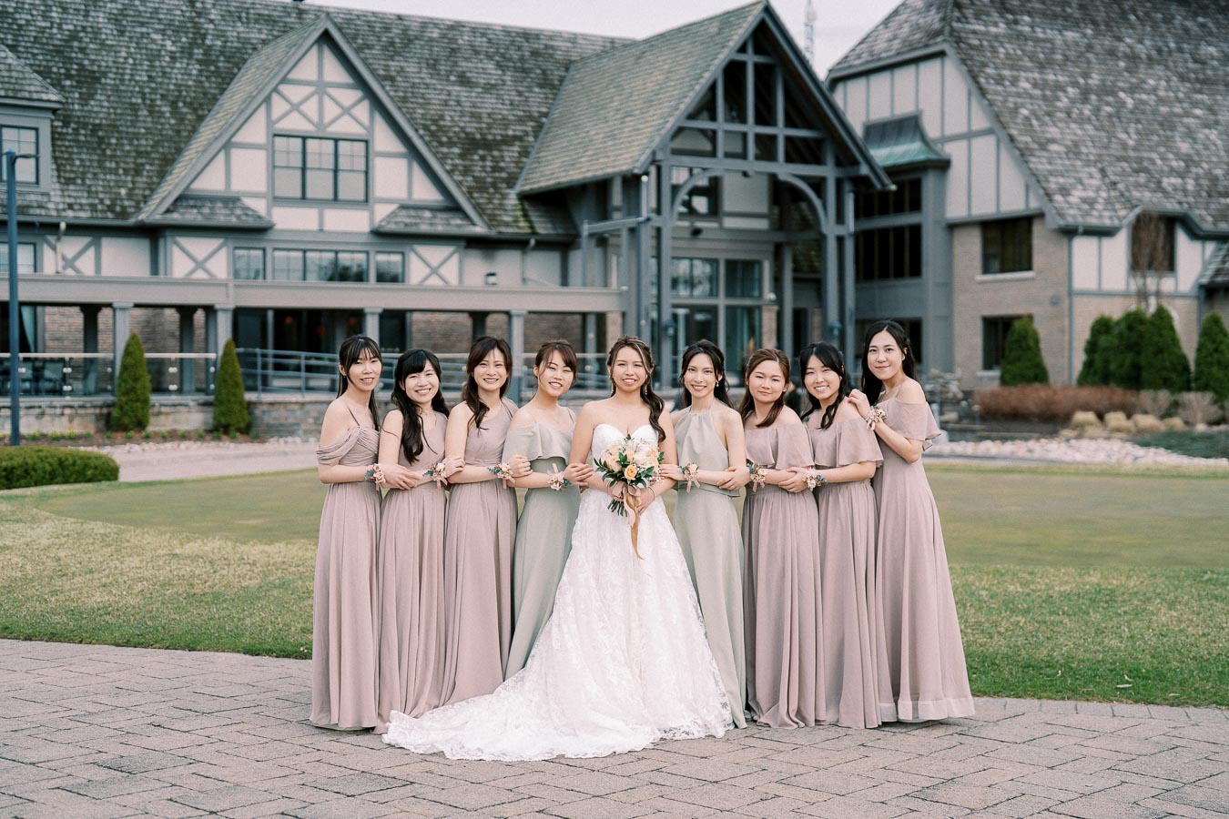 Wedding party posing in front of a Tudor-style building, with a bride in a white gown surrounded by bridesmaids wearing matching taupe dresses, standing on a paved area with a green lawn.