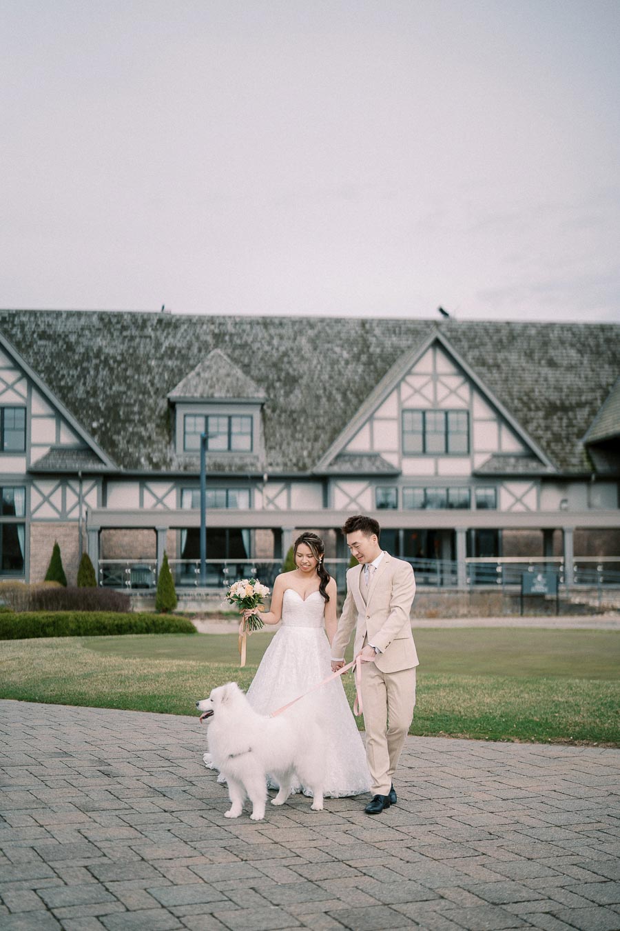 A bride and groom walk a fluffy white dog on a leash, smiling as they stroll outside a Tudor-style building, creating a charming wedding scene.