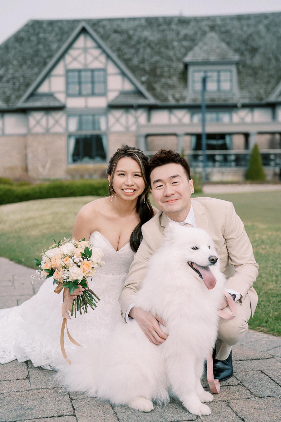 Bride and groom posing with fluffy white dog in front of a rustic building, showcasing wedding attire and bouquet.