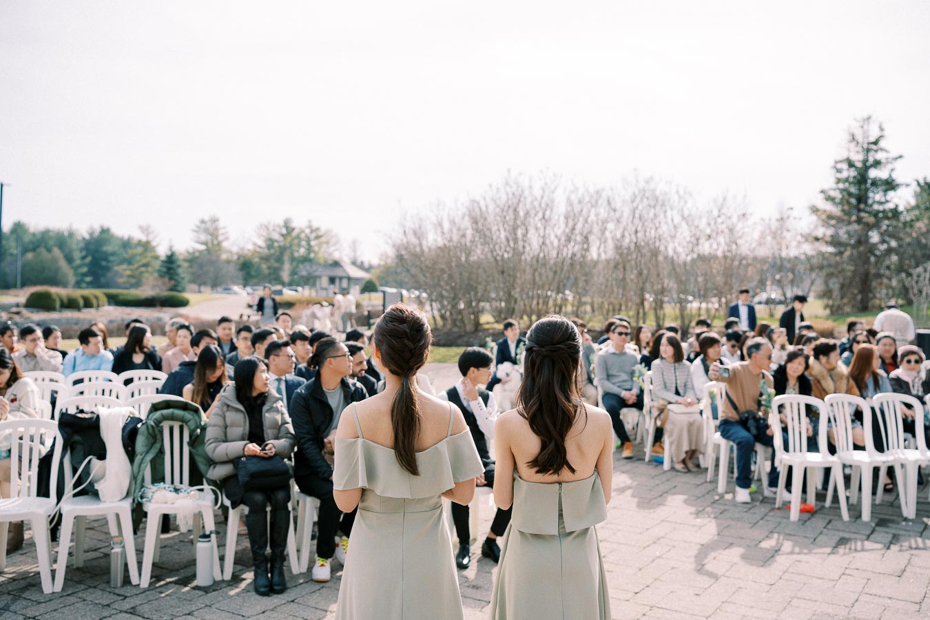 Two bridesmaids standing in front of a seated outdoor wedding audience, with bright sunlight and a scenic background of trees and open sky.