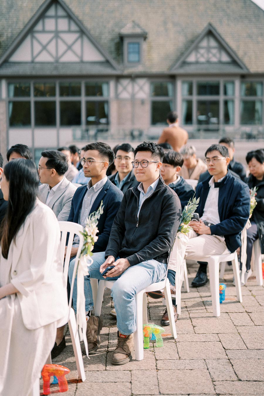 Audience seated at an outdoor event, with individuals dressed in semi-formal attire and a backdrop of a charming building.
