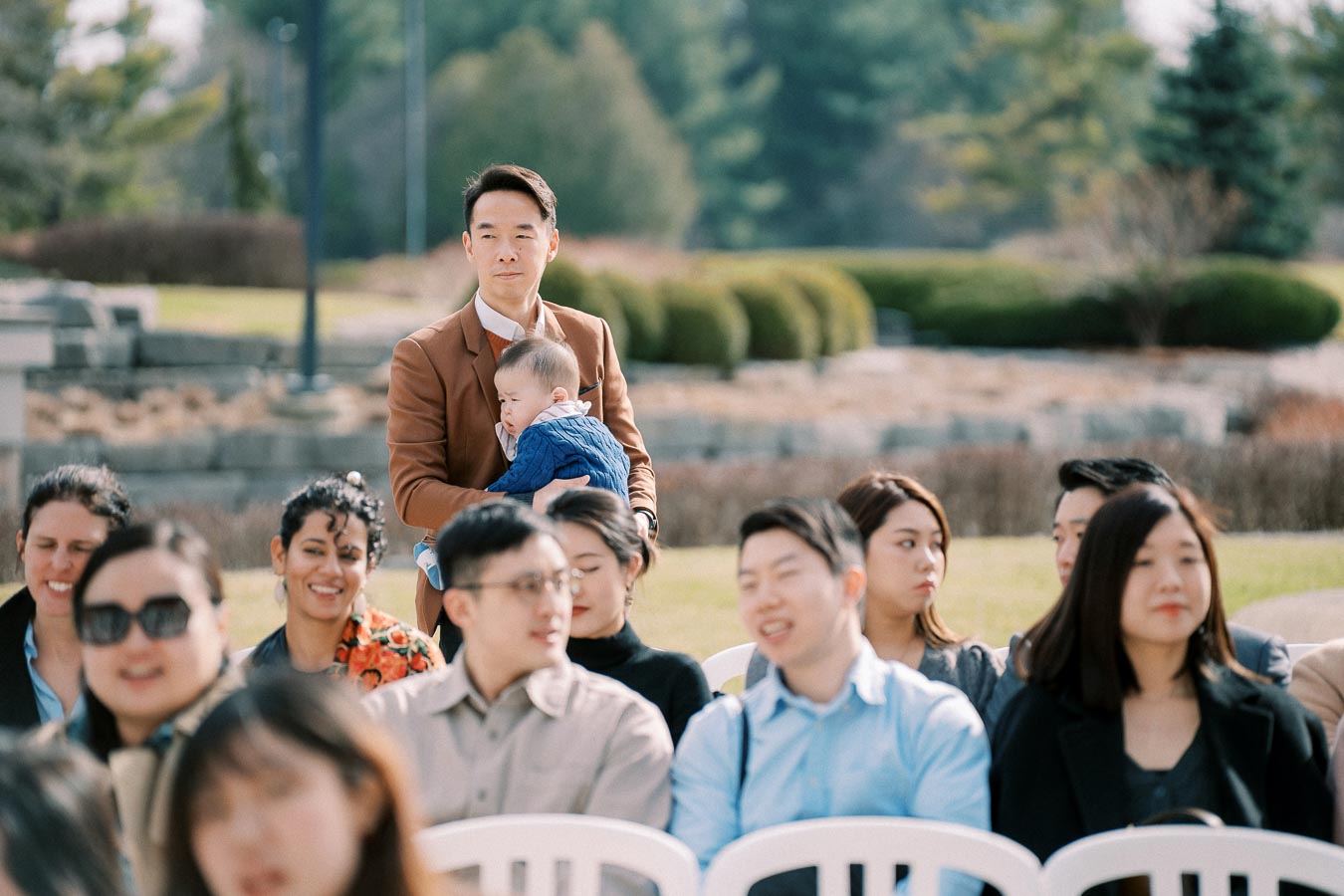 A diverse group of people sitting outdoors at a social event, with a man standing in the background holding a baby, surrounded by greenery and sunny weather.
