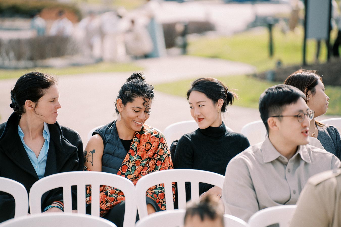 A group of people sitting on white chairs at an outdoor event, engaging in friendly conversation.
