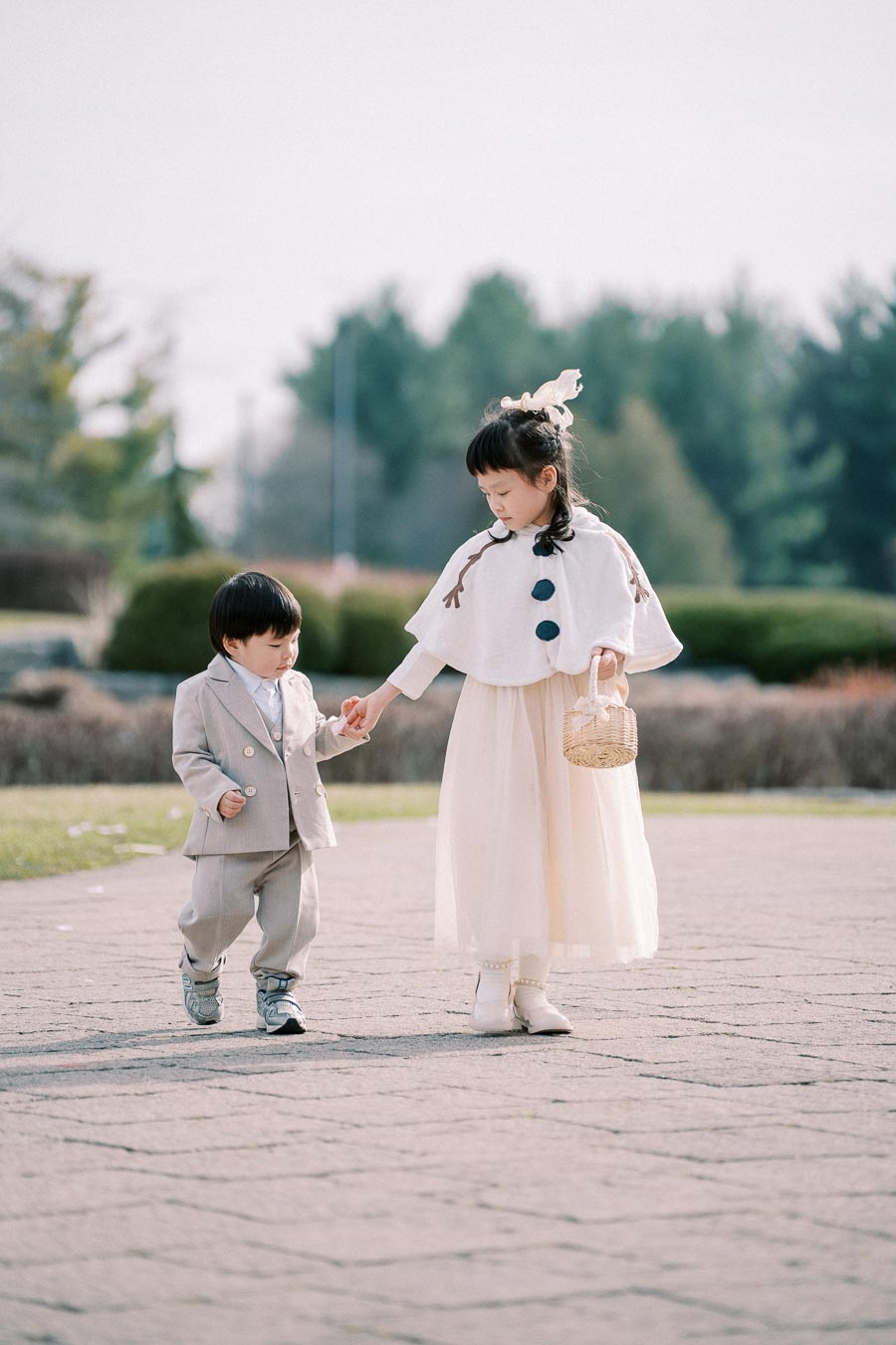 Two children dressed in elegant outfits walking hand in hand outdoors; the girl wears a white cape and skirt, holding a basket, while the boy is in a gray suit.