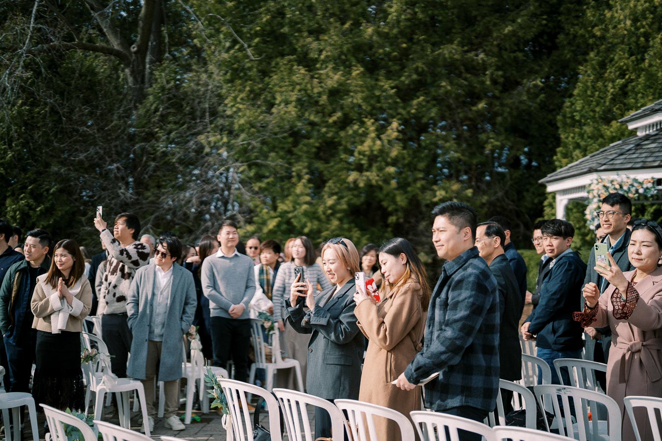 Outdoor wedding attendees stand and take photos, surrounded by greenery and a gazebo.